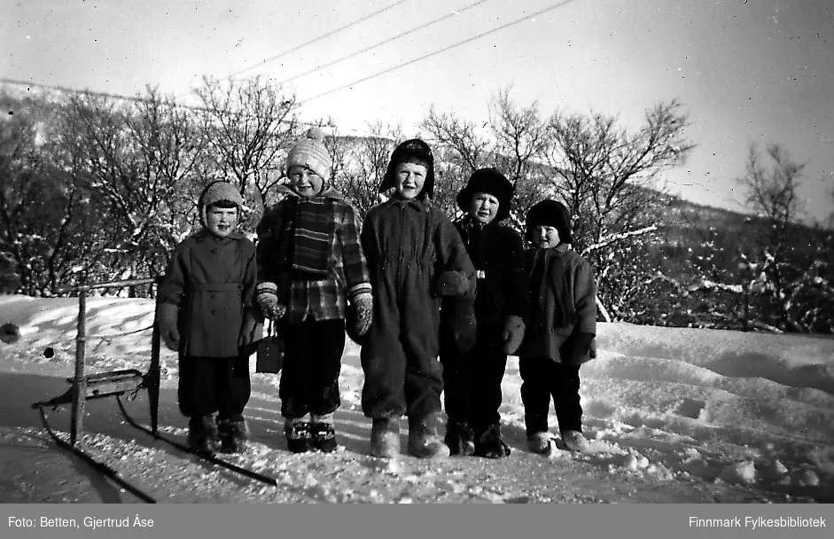 Fem barn poserer til fotografen på en veikant i en solig vinterdag i 1955/56. Jentene og guttene er kledd i vinterjakker, luer og votter. Noen av dem har komager på seg. Ved sidan av dem står en spark. Fra venstre: Åstrid Betten, Bjørn Ottar Betten, Oddvar, Kjell og Torill Betten. Åshild, Oddvar og Kjell er søsken. Alle unger er søskenbarn til hverandre.