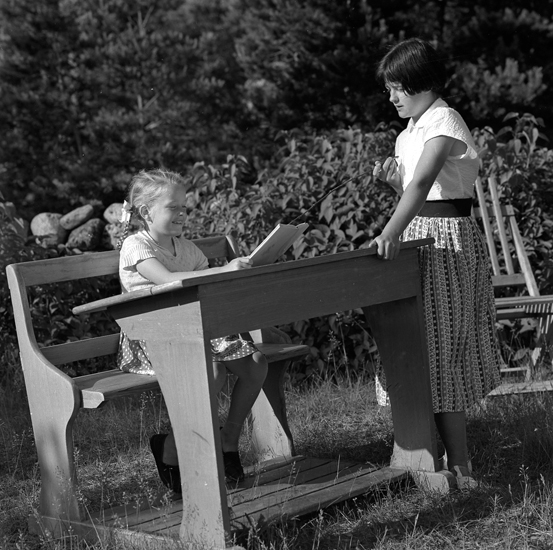 Ett barn sitter i en bänk som står utomhus, bredvid bänken står en annan person, möjligen en lärare.