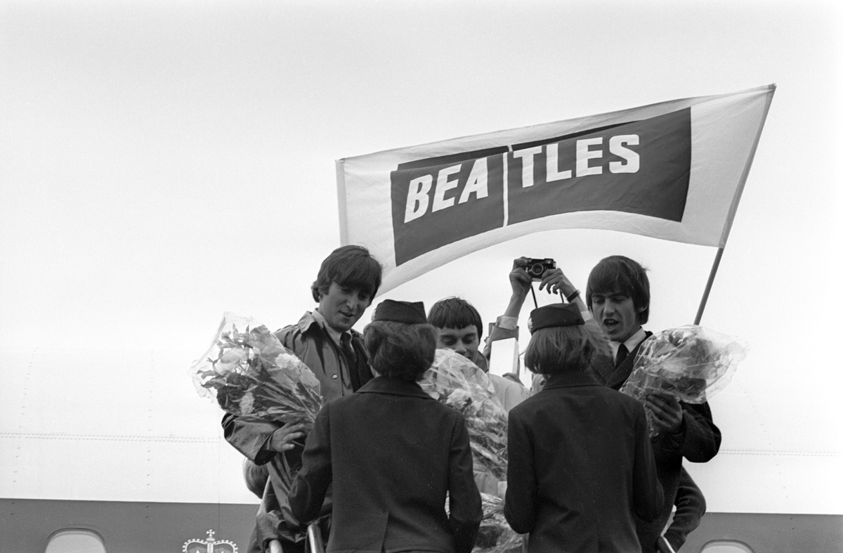 Det engelske bandet The Beatles skal ha konsert i København. Fra venstre John Lennon, Jimmy Nicol og George Harrison snakker med flyvertinnene på flytrappa. Popgruppa fløy med flyselskapet BEA (British European Airways) derav plakaten.
