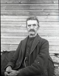 Portrett av en eldre mann sittende foran en husvegg. Mannen 