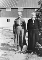 To eldre kvinner ute på en gårdsplass, driftsbygning i bakgr