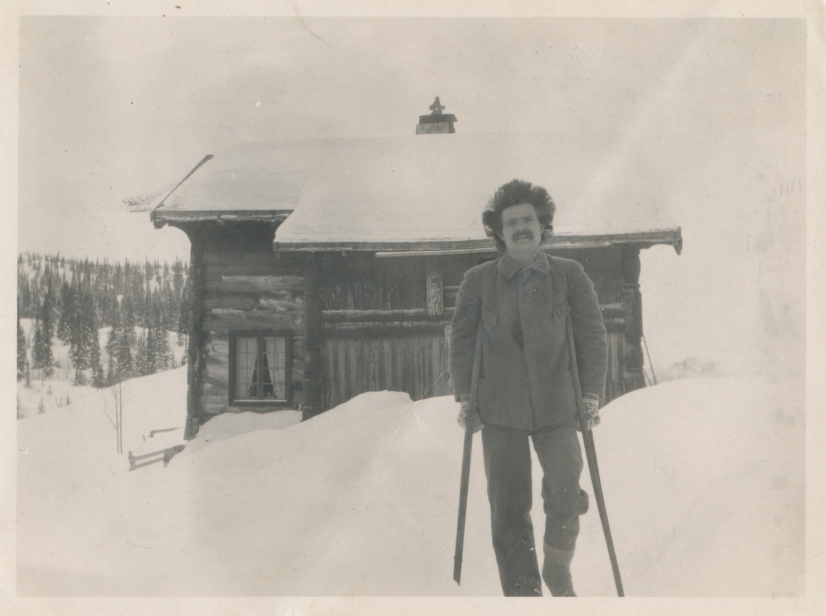 Mann med krykker foran ei hytte i vinterlandskap. Skjeldrup som ung.