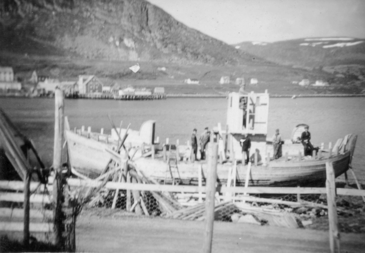 Flere menn på dekket til en båt som står på land i fjæra i Kvalsund sentrum før evakueringa. Båten kan være "Brødrene" som senere fikk navnet "Steinfjell" og en av eierene var Theodor Fredriksen. Personene på bildet er ukjent.
