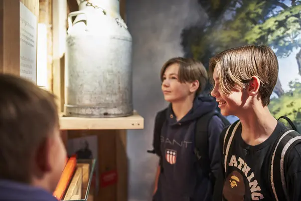 Tre unge gutter (elever) står og ser på et melkespann, bak dem er et maleri av et eiketre (eika)