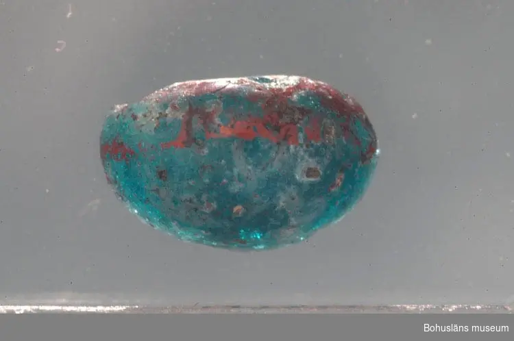 Del av glaspärla, halv. Pärlan är blå/turkos och har en röd dekorlinje. Tillverkningsavfall. 

Föremålet är placerat i back märkt med "övrigt material".