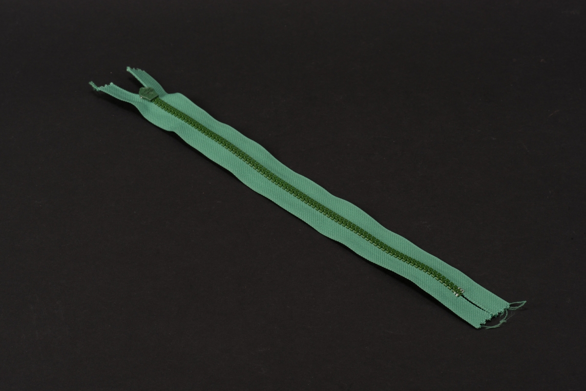 Grön dragkedja med textilband, häktor av plast och löpare och dragtapp av metall. På dragtappen inpräglat GUSUM.