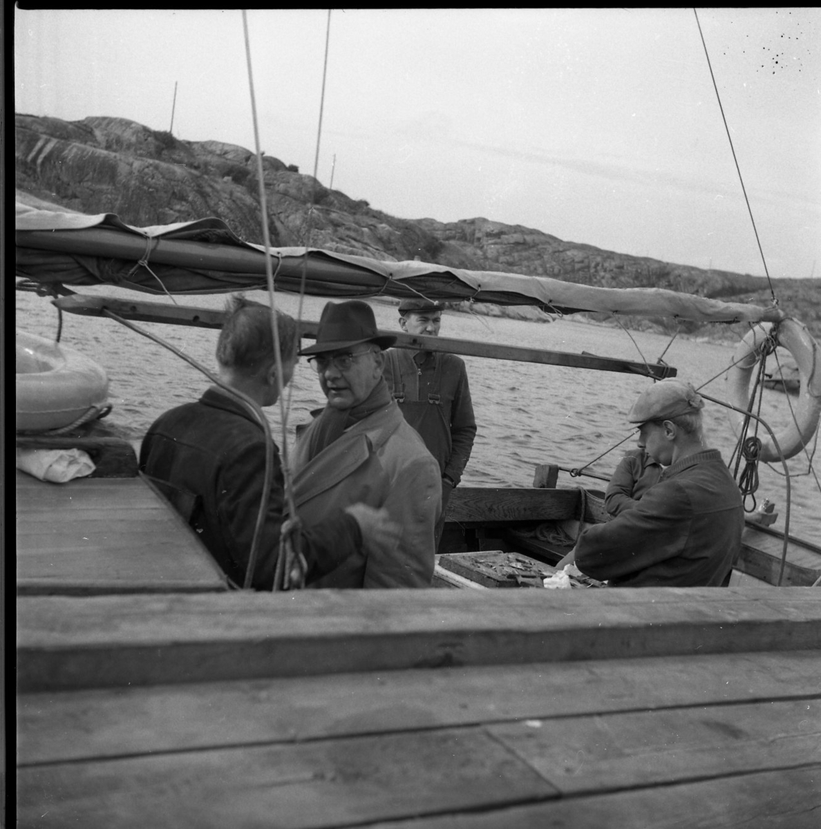 Fem män i en träbåt med mast och inombordare som ligger vid en brygga. En man i hatt och ljus rock är möjligen Osse Gustafsson.