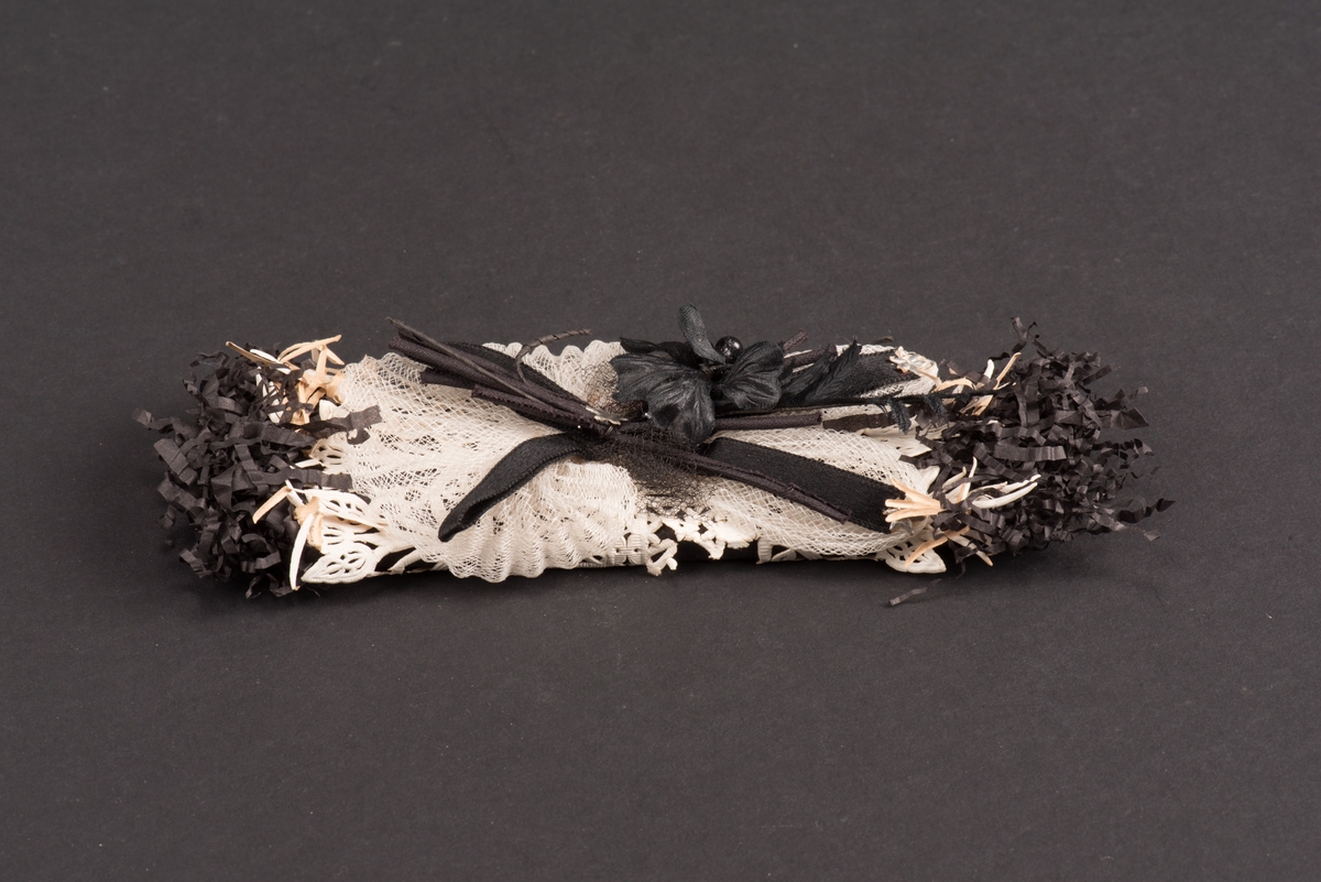 Platt begravningskaramell inlagd i svart silkespapper med klippta och krusade ändar. Omlindad med ett vitt perforerat papper. På ovansidan ligger en blomma med en rosett i sidenband på en bit vitt stärkt tyll.