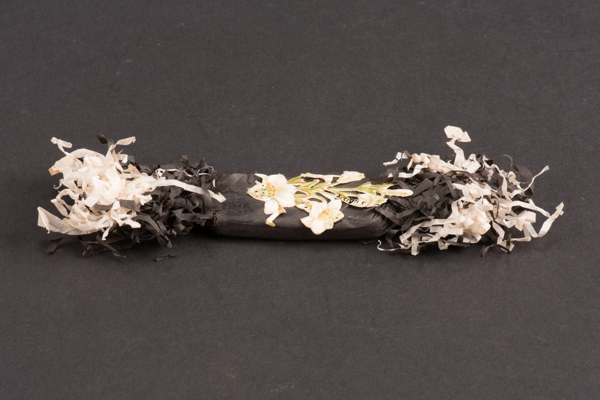 Platt begravningskaramell inlagd i svart silkesppapper med klippta och krusade fransar i svart och vitt i bägge ändarna.
På ovansidan dekorerad med ett påklistrat bokmärke i form av liljor.