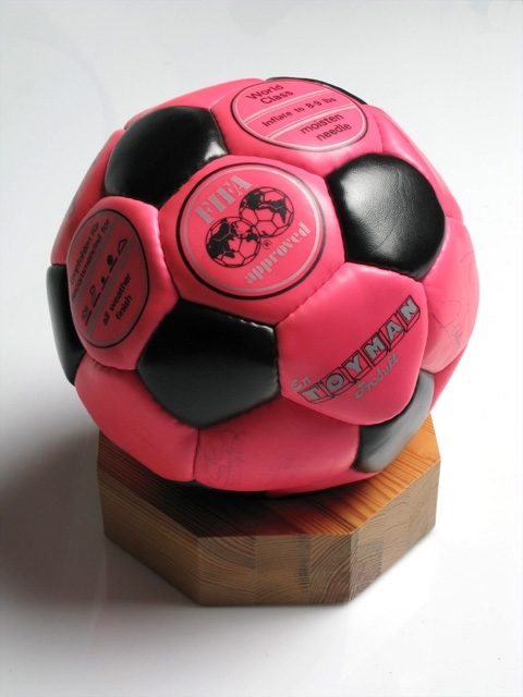Rosa fotboll med svarta femkantiga inslag. Fotbollen har FIFA:s
logotyp  och en del utländska fotbollsspelares autografer.
Övrig text på fotbollen World Class, Inflate to 8-9 lbs, moisten needle, all weather finish.

Träsockel, åttakantig, i ljust trä.