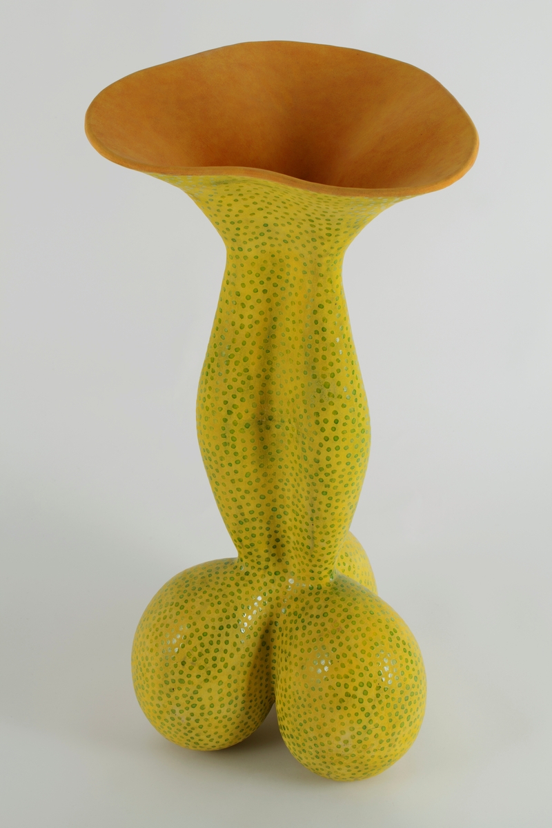 Stor, skulpturell krukke i leirgods. Organisk form som minner om en blomst; nederst tre belgformer, og opp fra denne en svulmende korpus med buede vegger og vid, bølgende munning. Formuttrykket er samtidig kroppslig og vitalt som en muskel. Oransje begitning innvendig, gul med grønne delvis transparente runde glasurflekker utvendig.