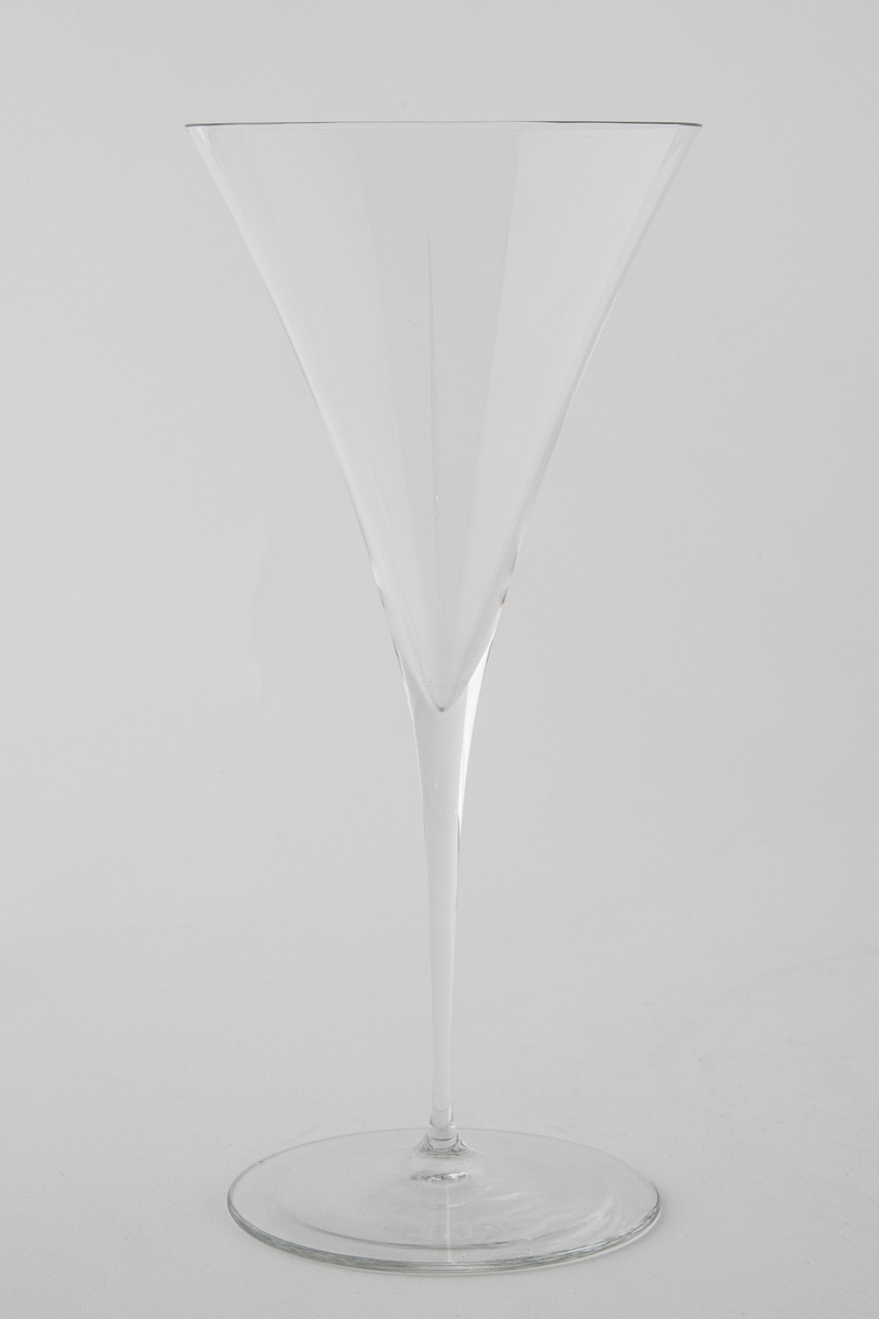 Høyreist vinglass i klart glass. Konisk kupa som glir over i en tynn stett, som hviler på en sirkulær fotplate.