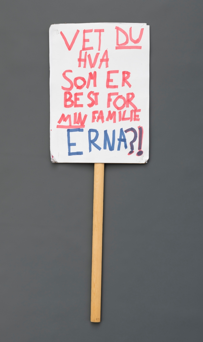 Håndtegnet to-sidig plakat med trepinne/-håndtak. På ene siden rødt kvinnetegn og bokstavene SU. På andre siden teksten: "Vet du hva som er best for min familie, Erna?! Hvitt papir med rød, sort og blå tusj.