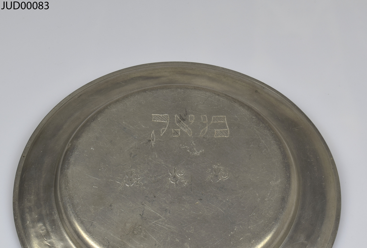 Tallrik tillverkad av tenn. Dekorerad med punsad dekor i form av hebreisk text längs med kanten och stiliserad blomma på spegeln. Hebreiska bokstäver inristade på baksidan.