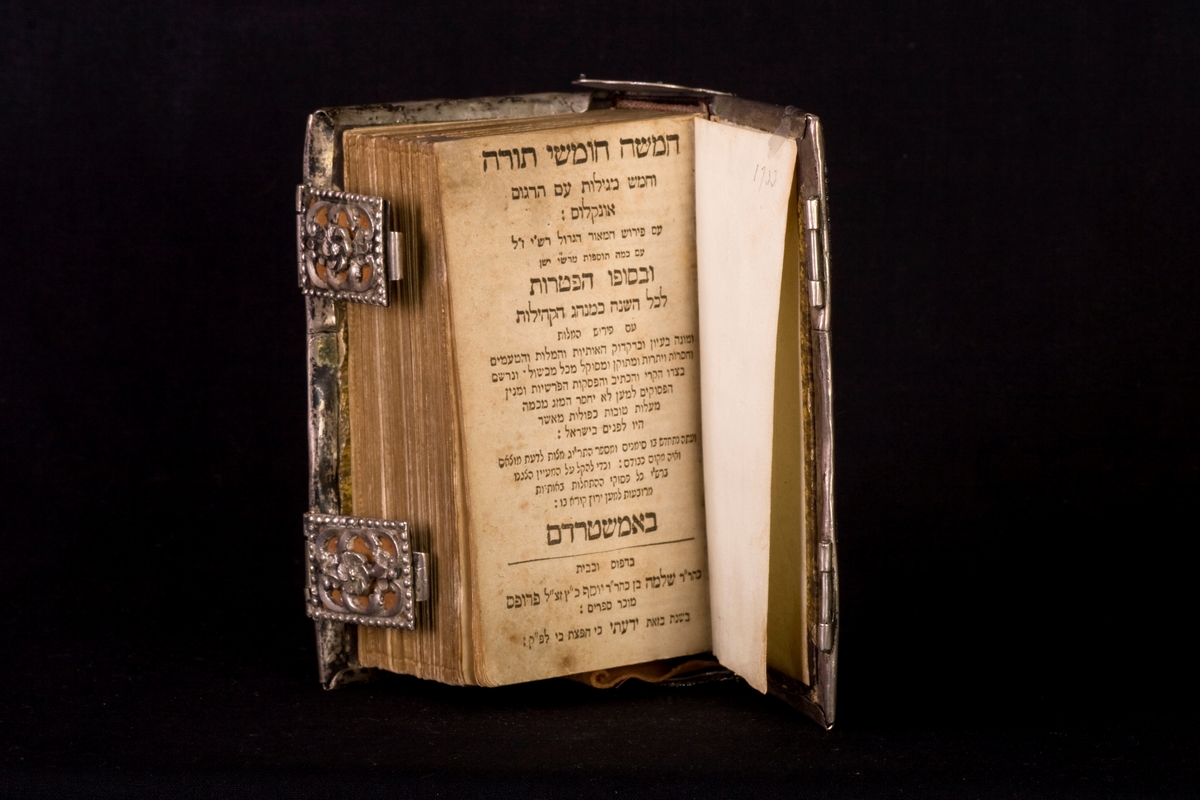 Siddur med sidor av papper, omslag av silverfiligranarbete och dubbla låsspännen. Skriven på hebreiska. Handskriven text på pärmens insida.