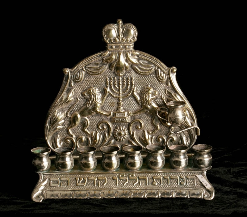 Chanukaljusstake tillverkad av silveröverdragen mässing. Dekorerad med en krona, hebreisk text samt en menora flankerad av två lejon. Bakstycket är fastsatt mot botten med skruvar i bakkant.