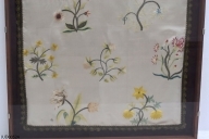Del av en gardin tillverkad av vitt siden. Fransar av koppartråd längsmed kanten. Dekorerad med broderade blommor i olika färger och utformning. Monterad i glas och ram.