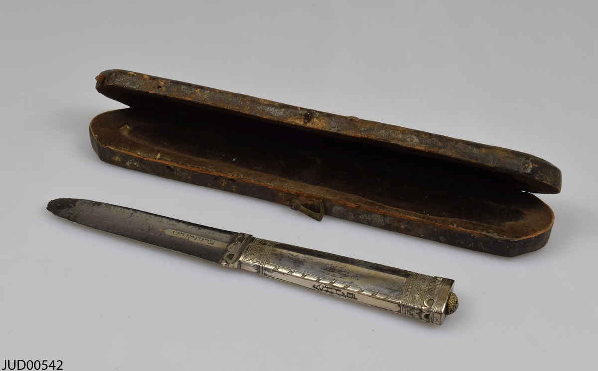 Omskärelsekniv med tillhörande etui. Kniven är tillverkad av silver och är dekorerad med ristade blommor. Etuiet är tillverkat av trä och därefter klätt utvändigt med läder, och invändigt med brun sammet.