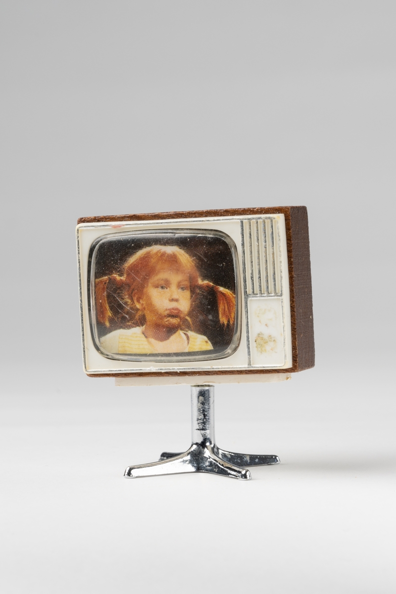 TV till dockskåp, av brunt trä, vit plast på fram- och baksida. Fot av silverfärgad plast. I rutan färgfoto av Pippi Långstrump.