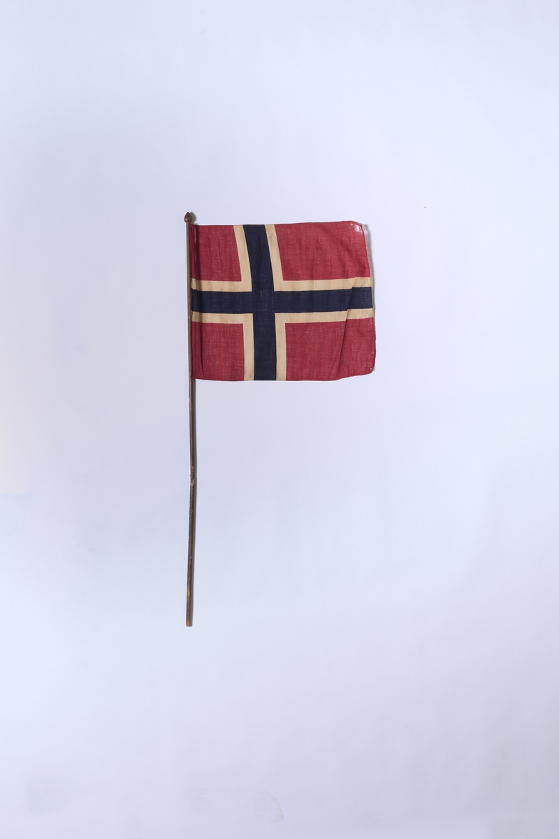 Det norske flagg, montert på trestang. Flagget er i bomull og har trykte farger. Stanga ser ut til å ha brukket for så å bli reparert. 