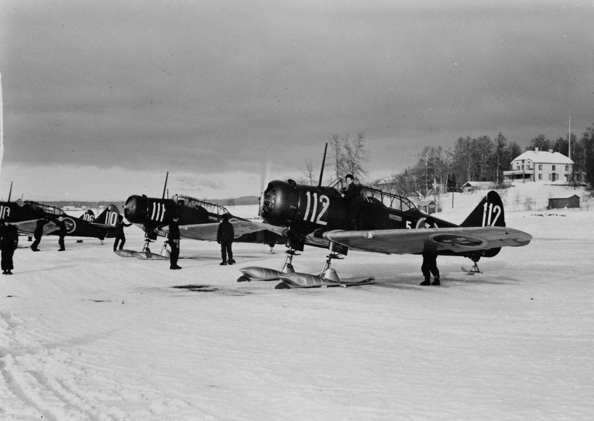 Flygplan SK 16A utrustade med skidor står på linje, vintertid. Flygplansmekaniker i arbete. Flygplan tillhörande Krigsflygskolan F 5 och märkta nummer 112, 111, 110, 106.