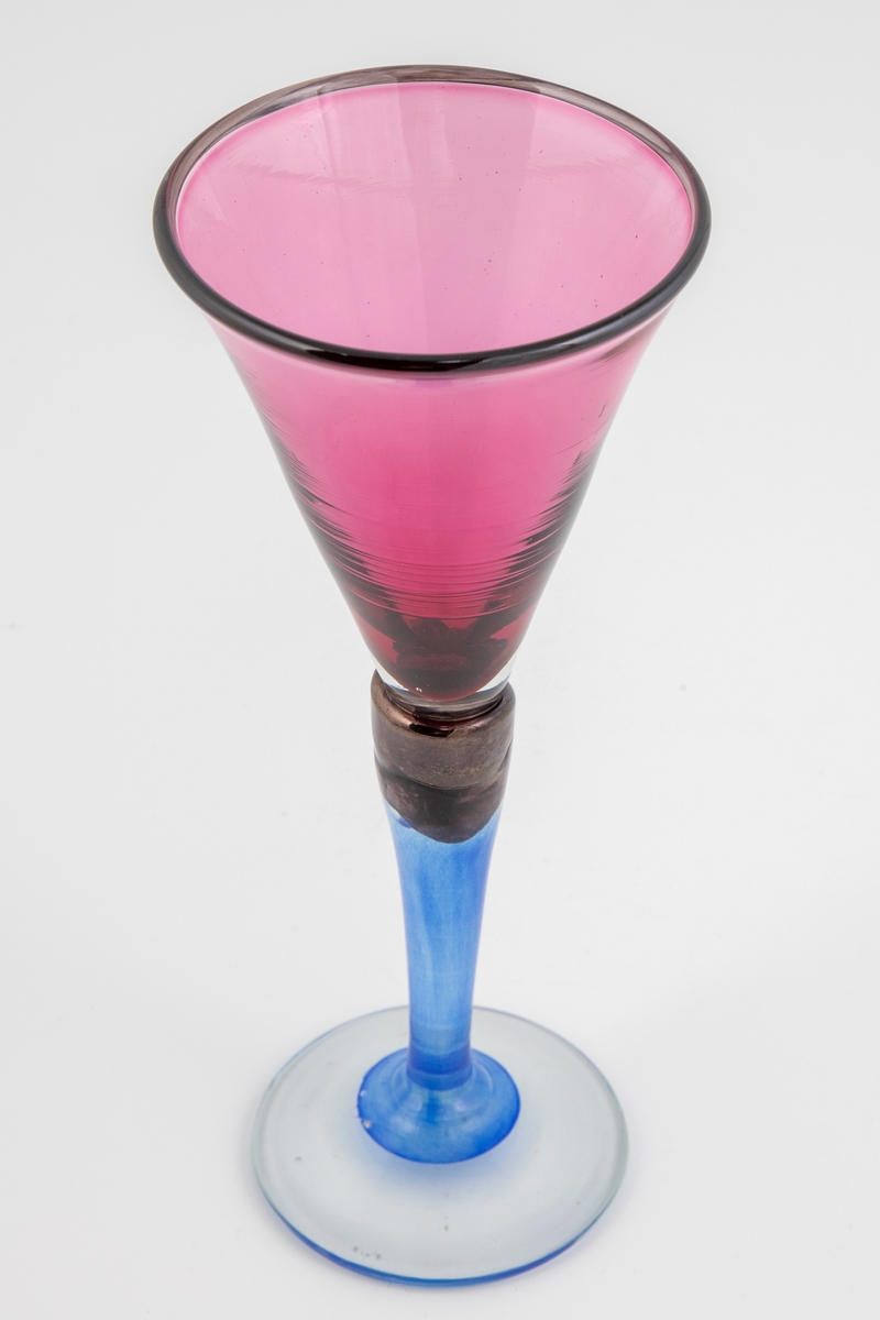 Vinglass med lillafarget konisk kupa, som hviler på en stett i blålig glass. Munningen er markert med mørkt glass. Foten er svakt buet og i klart glass.