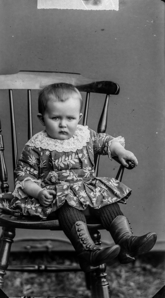 Jente antatt 2 år som sitter på pinnestol, kledd i blomstret kjole med blondekrage og høye skinnsko.