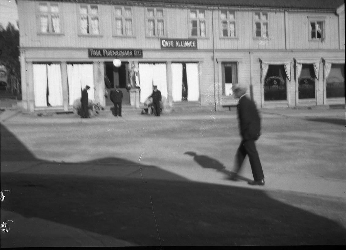 Stadsbild från Tromsö. På en husfasad skyltar för "Paul Figenschaus Eftr." och "Café Alliance". En man går över gatan.