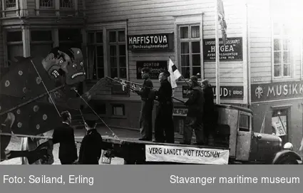 Antageligvis 1. maitog i Stavanger sentrum, 1939. Fire ungdommer på lasteplanet til en lastebil. På bilen står det "Verg landet mot facismen". Bildet er tatt utenfor "Kaffistova", som la rett ved siden av torget i Stavanger.