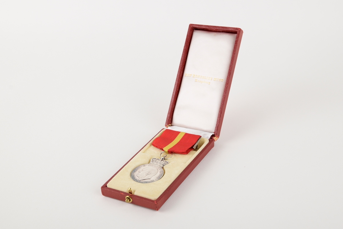 Et eksemplar av "Kongens fortjenstmedalje" i originalt etui. 

Sirkulær medalje med ordensbånd, oppbevart i rektangulært etui. Etuiet har motiv av en krone i sølv på lokket.
