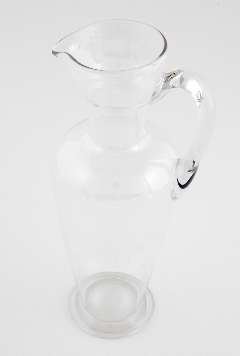Balusterformet mugge med nebb i klart glass. Påsatt hank samt sirkulær fotring.