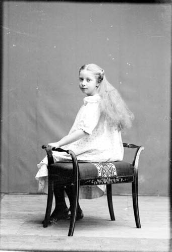 En flicka i ljus klänning och lång hår sitter på pall. Fotografi snett bakifrån.