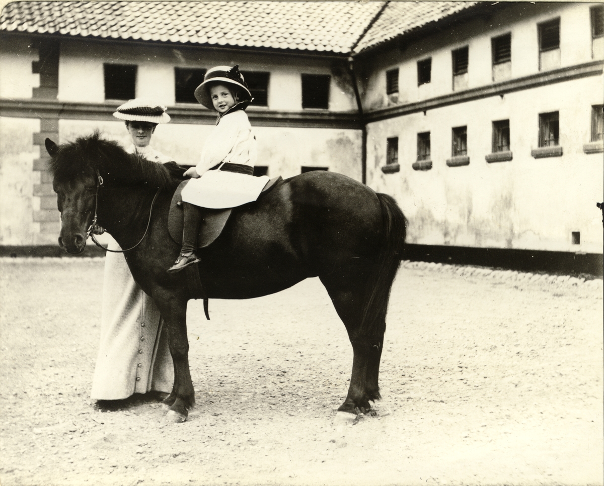 Mimi Egeberg rir på islandshesten "Dokka" på gårdsplassen til Bogstad gård. Moren Nini Egeberg holder hesten. Fotografert 1911.

