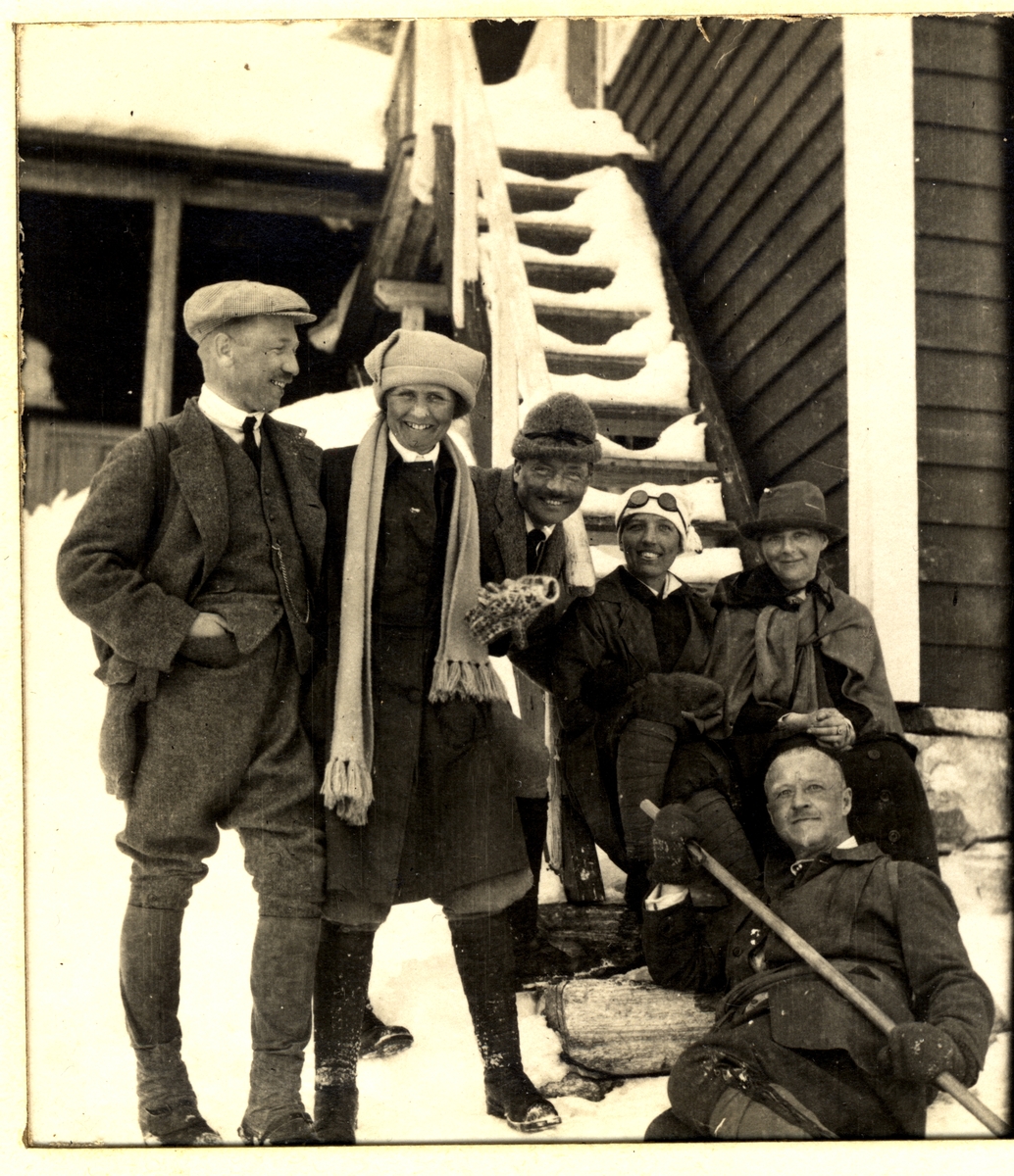 Medlemmer av skiklubben Fram med fruer på skiferie på  Finse. Fra venstre Westye P. Egeberg, og Agnes Løvenskiold og Fritz Treschow,. En av kvinnene på trappen er Lizzie Beer. Fotografert mars 1916.
