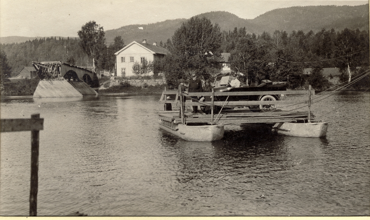 Bilen til Westye Egeberg trekkes over Sveen sund(?) på enkel taubåt bygget over to småbåter. Sjåføren sitter i bilen, Westye Egeberg og søsteren Evelyn står foran. Til venstre ses restene av en bro ved et bolighus. Fotografert 1916.