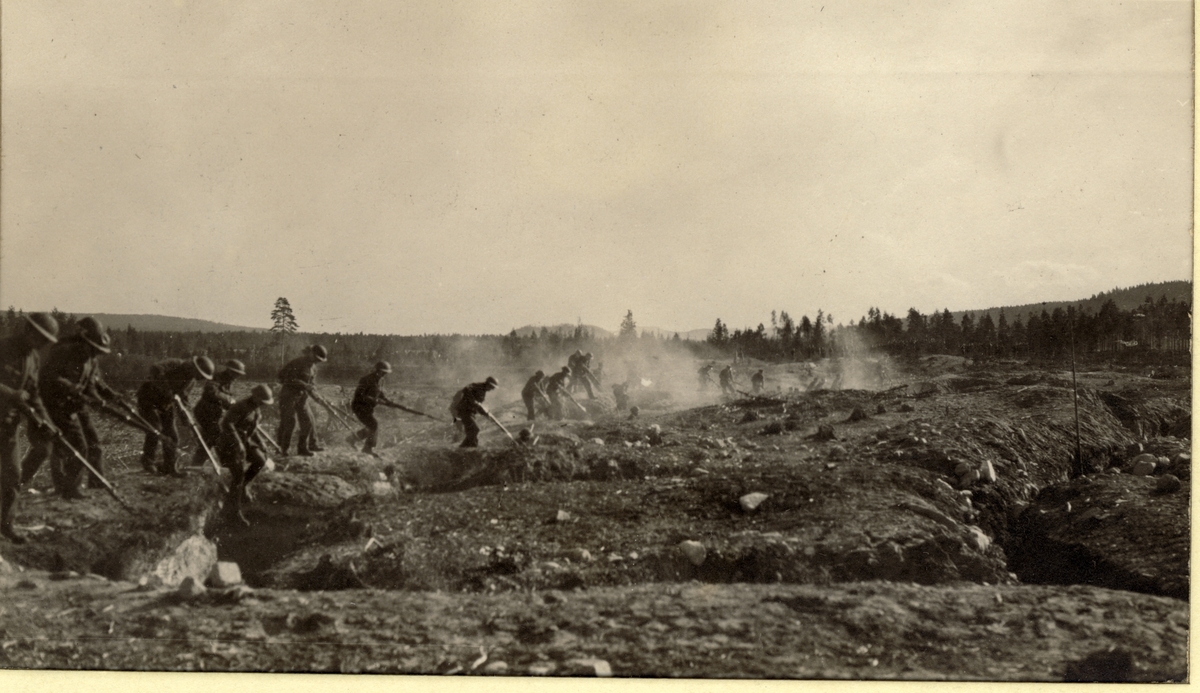 Infanterister på militærøvelse gjenskaper «Slaget i Flandern» fra første verdenskrig. Her erobres fiendens skyttergraver. Øvelsen skjer antagelig ved Infanteriets Skytterskole på Terningmoen i Elverum. Fotografert 1918.