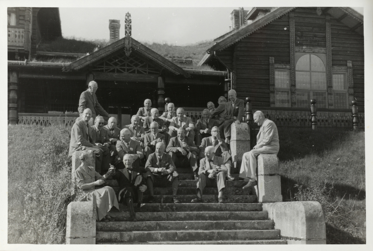 Medlemmer av skiklubben Fram samt et par kvinner samlet på trappen opp til Fjeldheim gård, opprinnelig jakthytte, ved Prestbuvatnet, Meldal. Fjeldheim gård er nå en del av Orkla Industrimuseum. Fotografert 1952.