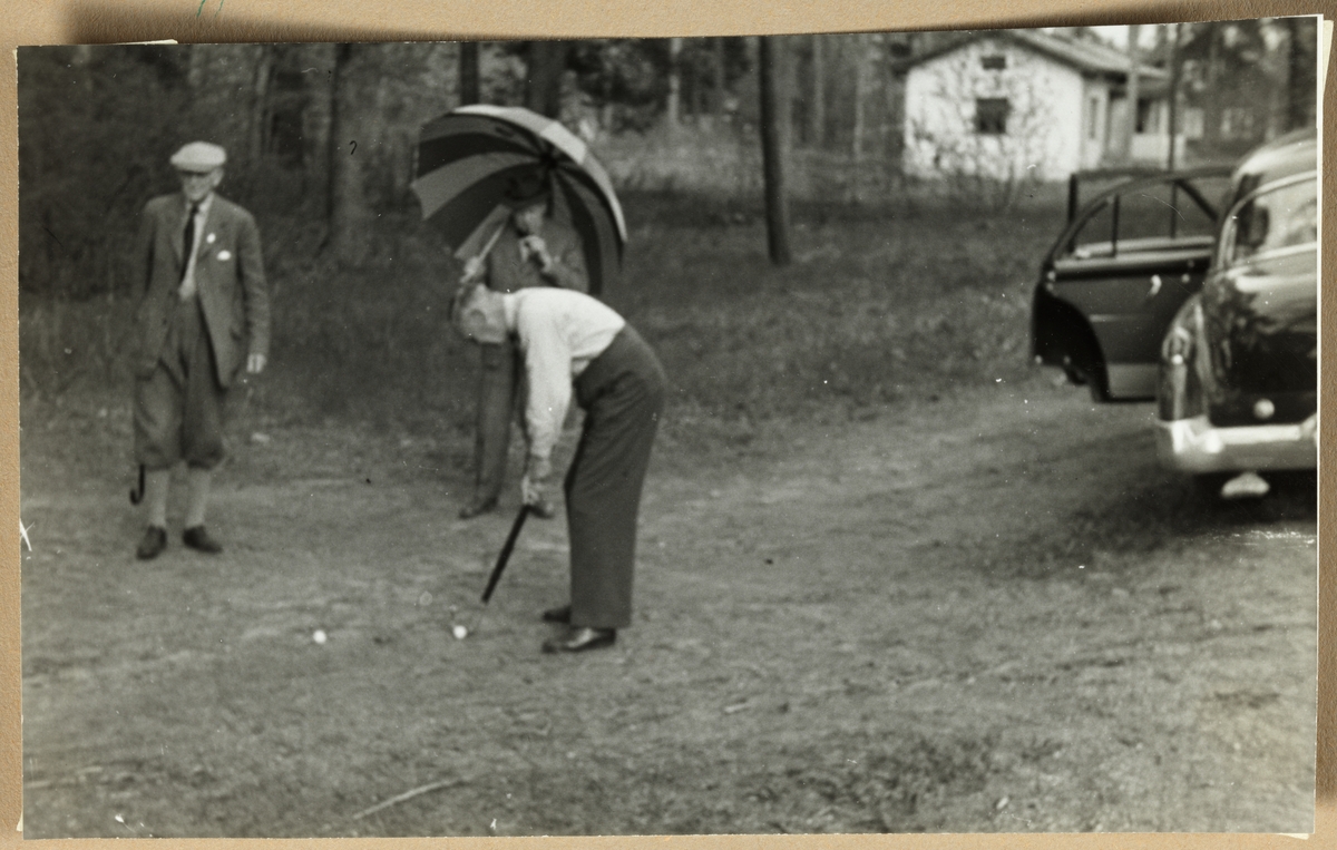 Tre menn fordriver tiden med noen slag golf mens de venter på flere deltakere. Til venstre Aksel Hansen og til høyre August Walle Hansen(?). I midten Kjell(?) med parasoll/paraply. Antagelig fotografert i nærheten av Dejefors, Sverige, mens skiklubben Fram var på besøk der i mai 1954.