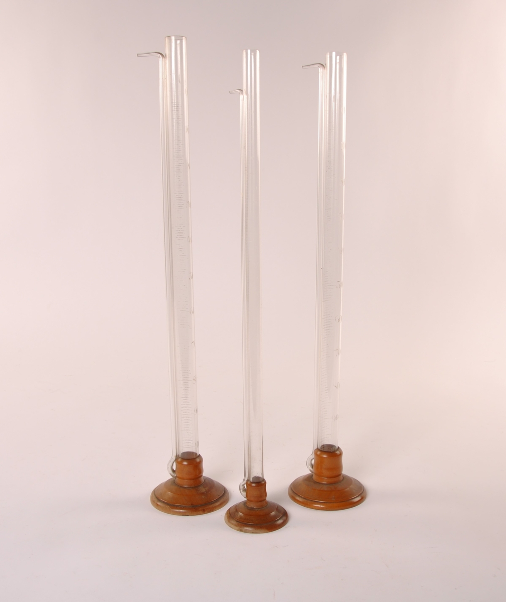 3 stk sylindriske målebegre i klart glass. Med fot i dreid tre. Tynn sylinder ved siden av hoved sylinderen.