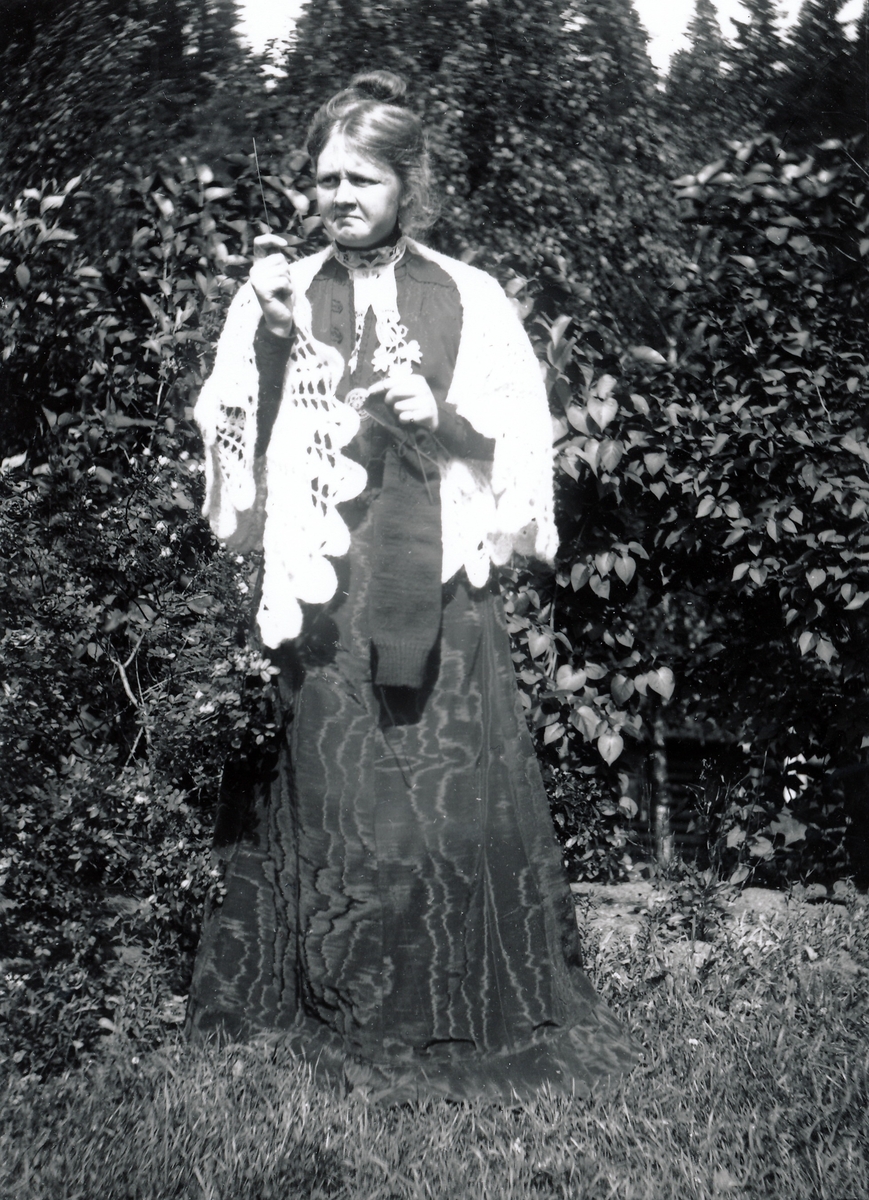 Uidentifisert kvinne stående blant busker i hage. I bakgrunnen er det syriner. Kvinnen er iført lang taftkjole og  lyst, heklet sjal over skuldrene. Oppsatt hår, holder strikketøy (strømpe) i hendene.