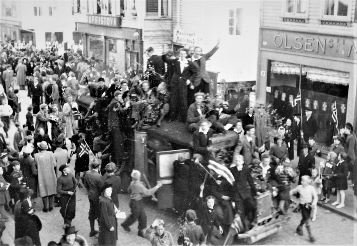Feiring av freden i Haugesund 8.mai 1945. Større folkemengde i Haraldsgata. Midt i bildet en buss med jublende personer rundt og på taket av bussen. Norske flagg. Butikkrekke i bakgrunnen, med "Kaffistova" til venstre og "Olsens manufaktur" til høyre.