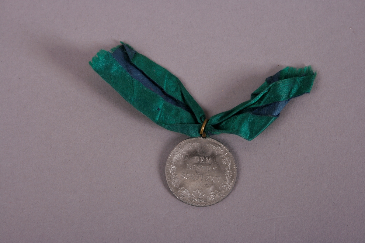 Medalje med grønt bånd, festet i ring på toppen.