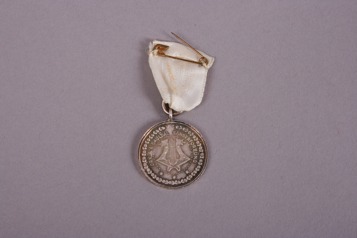 Medalje med hvitt bånd. Stevnet i Røros 1949 er skrevet rundt et portrett av en viking med bue. På baksiden en harpe med dragefjes ombranset av tekst og små blader. 
Teksten er: Orkla & Gauldal Sangerforbund.