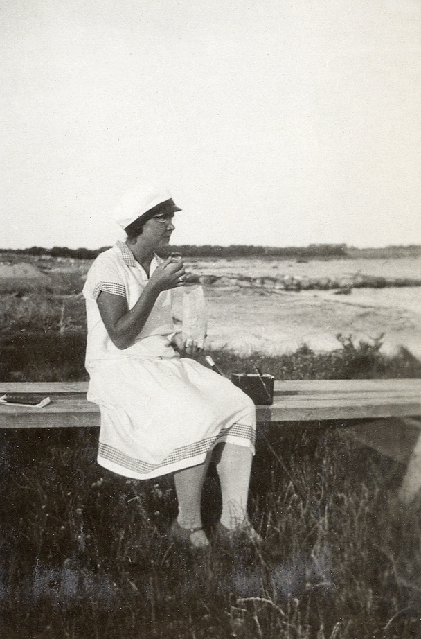 En kvinna i ljus klänning och studentmössa sitter och äter något ur en påse, vid en strandäng. 
Under fotot text: "Hällevik, 1929".