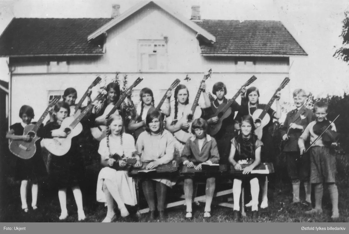Barnemusikken ved Julie og Kristian Kasbos søndagsskole i Spydeberg 1927. Musikantene er stilt opp for fotografen foran huset. 8 gitarister, 2 fiolinister (de eneste guttene) samt 4 piker foran som spiller på harpeleik(cither/zither).