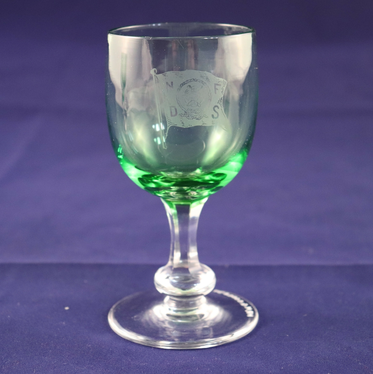 Hetvinsglass med sekskantet stett og rund plate nederst. Glasset er farget grønt med blank stett. Rederiflagget til Det Nordenfjeldske Dampskibsselskab er gravert inn på siden.