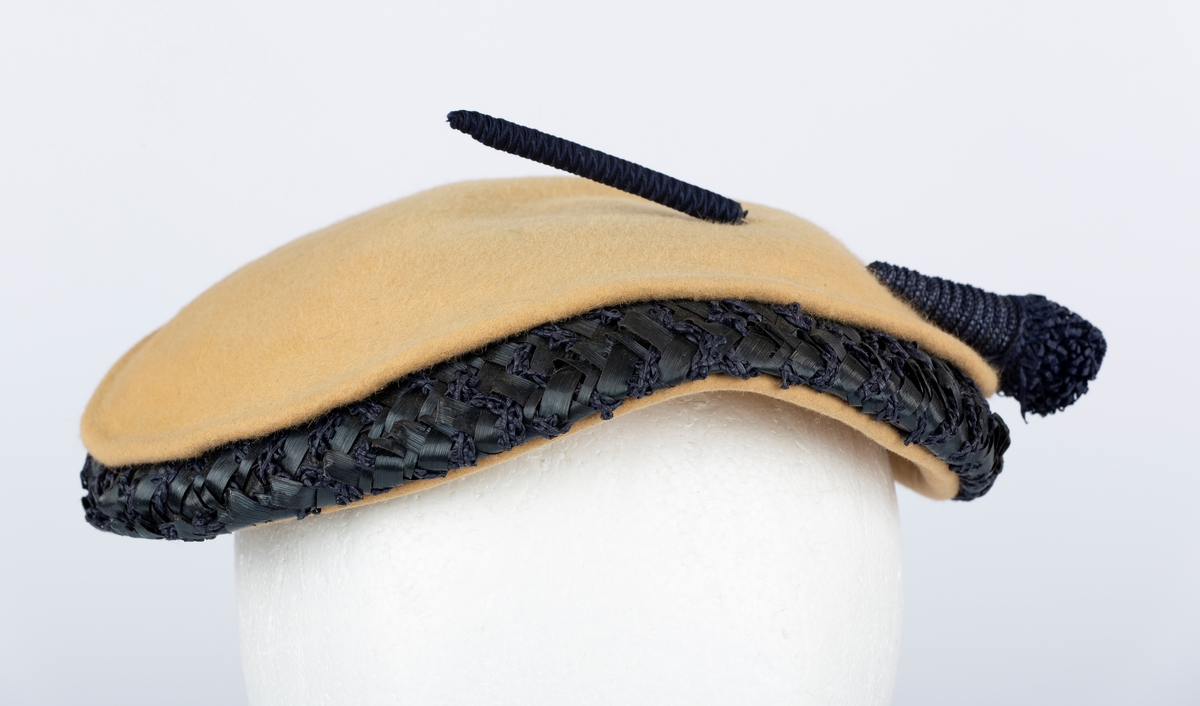 Flat liten hatt med kant av blå tråd og og bast.
Brukt av Olga Ambjørnrud under 2. verdenskrig