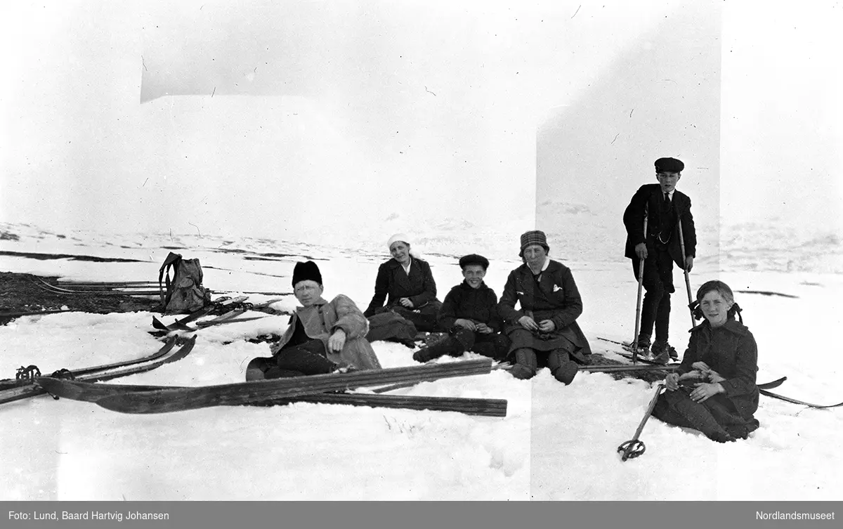 Gruppebilde, tre kvinner og tre menn på ski, sannsynligvis i Sulitjelmatraktene. Mannen til venstre er ukjent. Ungjenta til høyre er Signe Lund. Gutten som står ved siden av henne er Kristian Lund. Gjengen slapper av så både ski og staver ses rundt dem.