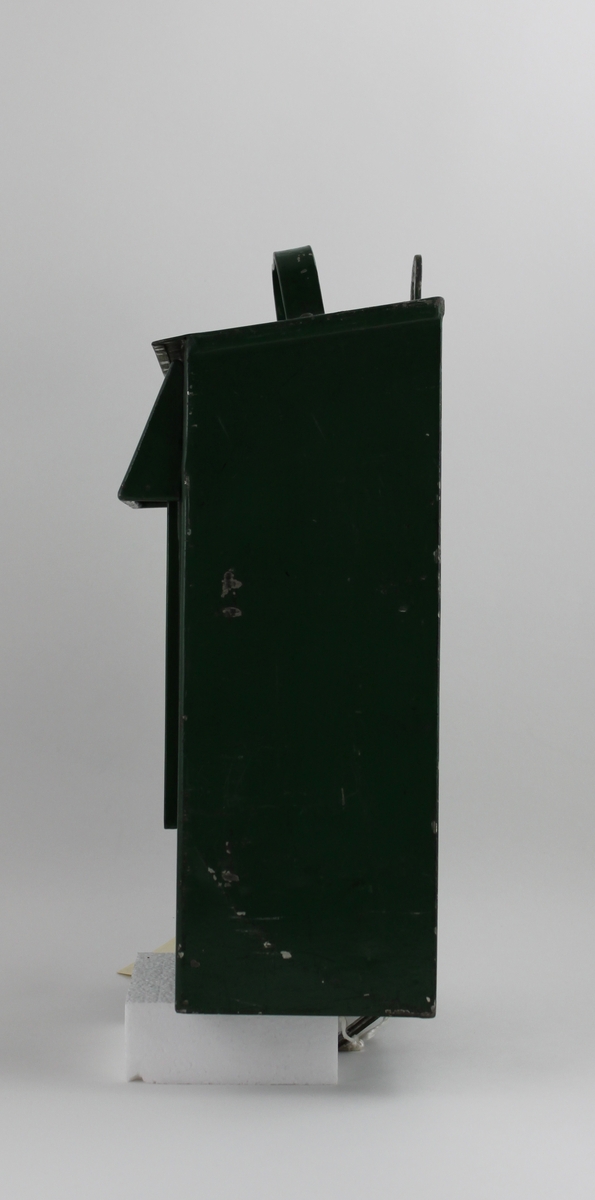 Brevlåda, med fronttömning, på klaffen över brevinkastet finns ett målat sigillförsett kuvert. På brevlådans tak finns ett handtag.