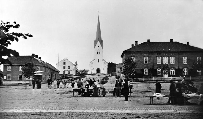 Svart-hvitt fot som viser et torg med handelsboder og torgkoner i "gammeldagse" klær, et hus i hver bildekant og Hamar domkirke bakerst i midten.